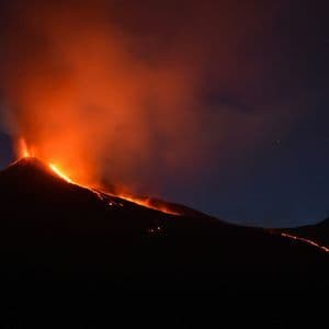 Un vulcano in eruzione di notte, con lava incandescente arancione che scende lungo il pendio e fumo che si innalza nel cielo stellato.