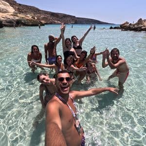 Un viaggio di gruppo WeRoad sorride per un selfie mentre si trova in acque limpide e turchesi in una cala rocciosa e soleggiata.