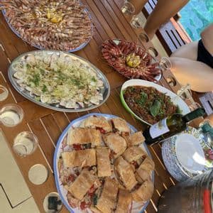 Un gruppo WeRoad in viaggio cena in barca, con una tavola imbandita di frutti di mare freschi, panini e vino bianco.