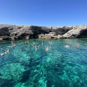 Un viaggio di gruppo WeRoad fa il bagno in acque limpide e turchesi vicino a una costa rocciosa sotto un cielo azzurro.