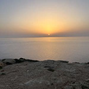 Il sole tramonta all'orizzonte su un mare calmo, visto da una scogliera rocciosa in primo piano.