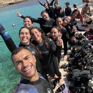 Un viaggio di gruppo WeRoad in mute sorride per un selfie di gruppo su un pontile vicino al mare, con l'attrezzatura da sub pronta.
