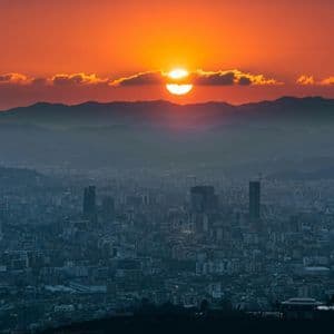 Un sole arancione intenso tramonta dietro una lontana catena montuosa, che si affaccia su una vasta città avvolta nella foschia.