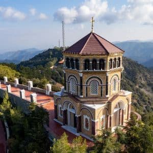 Une église ornée d'un toit de tuiles rouges est perchée sur un sommet boisé, dominant une vaste chaîne de montagnes sous un ciel partiellement nuageux.