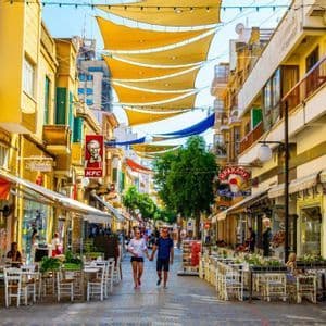 Un couple se promène dans une rue piétonne animée bordée de cafés et de boutiques, sous de grands stores de soleil jaunes suspendus entre les bâtiments.