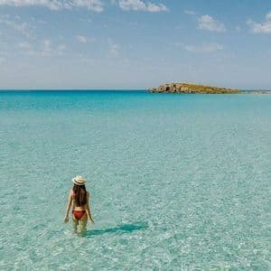 Une femme portant un chapeau de soleil et un bikini se tient dans une eau turquoise peu profonde et limpide, regardant vers une petite île à l'horizon.