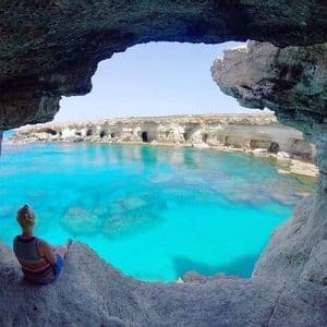 Une femme est assise à l'entrée d'une grotte marine, contemplant une baie calme aux eaux turquoise claires et des falaises rocheuses sous un ciel bleu.