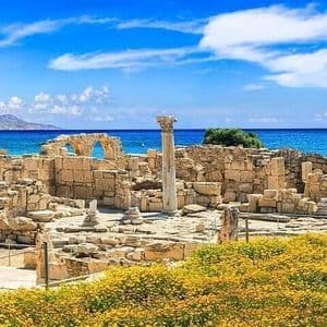 Anciennes ruines de pierre avec une colonne dressée, surplombant une mer d'un bleu éclatant, et des fleurs jaunes au premier plan.
