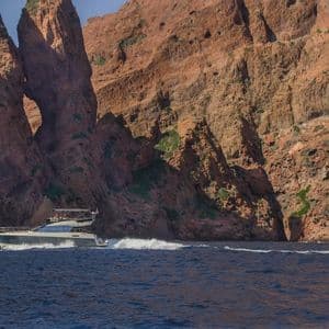 Un yacht à moteur navigue sur la mer d'un bleu profond le long d'une côte spectaculaire de falaises de roche rouge avec une arche naturelle.