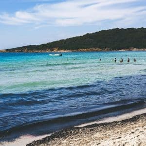 Des gens se baignant dans la mer turquoise d'une crique de sable, avec un petit bateau et une colline verdoyante en arrière-plan.