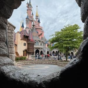 Un castello fiabesco rosa con torri blu e oro si ammira attraverso un'apertura in pietra grezza, che si affaccia su un cortile animato da persone.