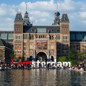 The iconic 'I amsterdam' sign in front of the Rijksmuseum, with people sitting around the edge of a large reflecting pool.
