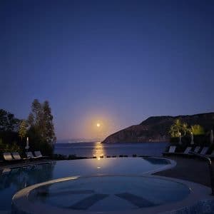 Una piscina di notte con vista sulla luna piena che si riflette sul mare calmo, vicino a una costa scura.
