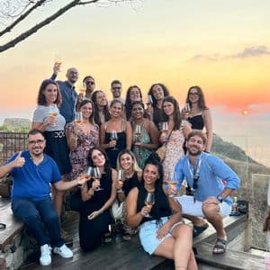 Un gruppo WeRoad in posa per una foto su una terrazza, con calici di vino, durante un tramonto dorato sul mare.