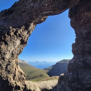 Un paesaggio costiero con verdi colline e isole lontane, ammirato attraverso un arco di roccia naturale in una giornata di sole.