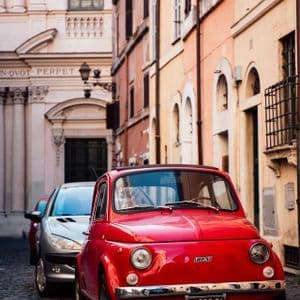 Un Fiat 500 rojo vintage aparcado en una estrecha calle empedrada con edificios históricos de fondo.