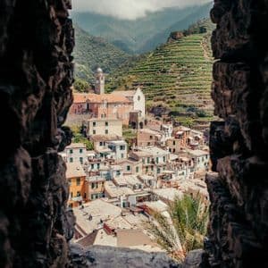 A view through a stone archway of a colorful hillside village with a church and terraced vineyards in the background.
