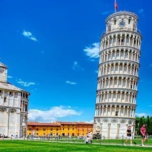 The Leaning Tower of Pisa stands on a green lawn next to a large cathedral under a bright blue sky, with people strolling nearby.