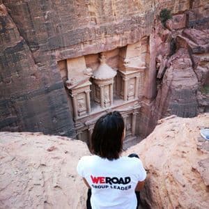 Un Group Leader WeRoad, di spalle, siede sul bordo di una scogliera rocciosa, ammirando un tempio scavato in un canyon di roccia rossa.