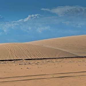 Vaste dune di sabbia dorata con tracce sui loro pendii sotto un cielo azzurro parzialmente nuvoloso.