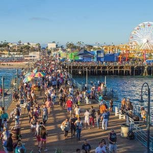 Una grande folla di persone cammina lungo un molo di legno soleggiato, con un parco divertimenti e una ruota panoramica visibili sullo sfondo.