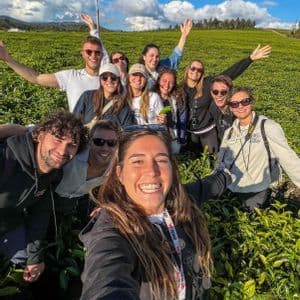 Un gruppo WeRoad in viaggio sorride per un selfie in una piantagione di tè verde, sotto un cielo azzurro e nuvole.