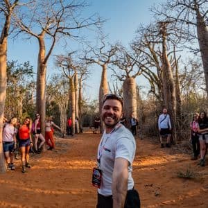 Un uomo sorridente si scatta un selfie con il suo gruppo WeRoad tra grandi baobab su un sentiero sterrato.