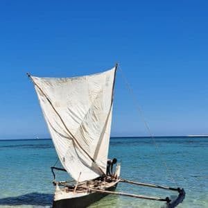 Una tradizionale barca a vela a bilanciere in legno con una grande vela bianca riposa in acque turchesi poco profonde vicino a una riva sabbiosa.