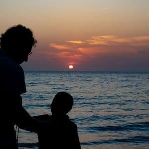 Una silhouette di un adulto e un bambino che ammirano il tramonto sull'oceano dalla spiaggia.