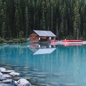 Una baita in legno si trova sulla riva di un lago calmo e turchese, con canoe rosse ormeggiate nelle vicinanze e una fitta pineta alle sue spalle.
