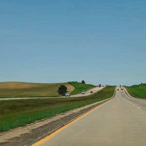 Un'autostrada attraversa un paesaggio di dolci colline verdi e marroni sotto un vasto cielo azzurro e limpido.