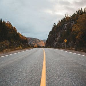 Una strada asfaltata deserta si snoda in una valle fiancheggiata da colline ricoperte di alberi autunnali sotto un cielo nuvoloso.