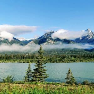 Un lago sereno è circondato da una pineta, con un'imponente catena montuosa coperta di nuvole che si innalza sullo sfondo sotto un cielo blu.