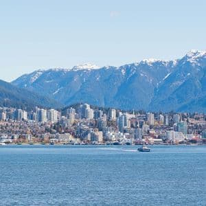 Uno skyline cittadino su uno specchio d'acqua, con grandi montagne innevate che si ergono sullo sfondo sotto un cielo sereno.