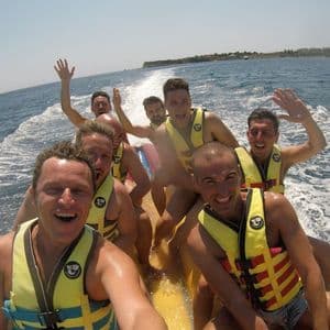 Eine WeRoad-Gruppe von Männern lacht und macht ein Selfie auf einem gelben Bananenboot im Meer.