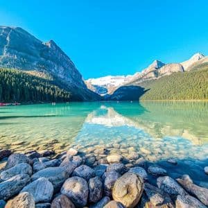 Un lago limpido e turchese con un primo piano roccioso è circondato da montagne ricoperte di pini e un ghiacciaio lontano sotto un cielo blu.