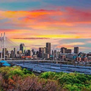 Vue urbaine avec pont à haubans et gare de triage, au-dessus d'arbres verts lors d'un coucher de soleil coloré.