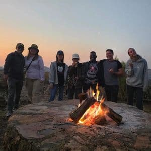 Un voyage en groupe WeRoad avec des gens souriants autour d'un feu de camp sur une colline au coucher du soleil.