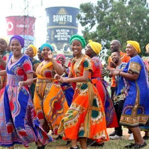 Un groupe de personnes vêtues de tenues africaines traditionnelles et colorées, dansant et chantant ensemble en plein air sur un terrain herbeux.