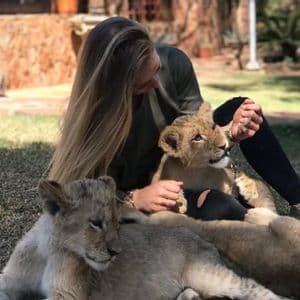 Une femme aux longs cheveux blonds est assise sur une pelouse et caresse deux jeunes lionceaux.