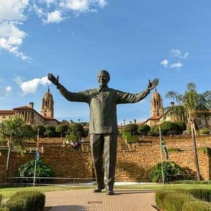 Une grande statue de bronze de Nelson Mandela se dresse, les bras tendus, dans un jardin devant un bâtiment majestueux et symétrique sous un ciel bleu.