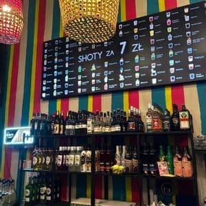 Eine bunte Bar mit Regalen voller Spirituosen und einer großen digitalen Menütafel, die eine Auswahl an Shots vor einer gestreiften Wand bewirbt.