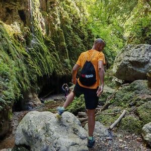 Ein Mann mit einem WeRoad-Rucksack wandert durch eine enge, moosbewachsene Schlucht und steigt über einen großen Felsen in einem Bach.
