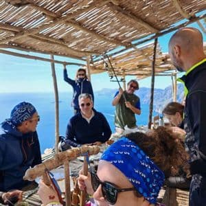 Un viaggio di gruppo WeRoad si riunisce attorno a un tavolo di legno sotto un tetto di paglia, con vista sul mare e sulle montagne costiere.