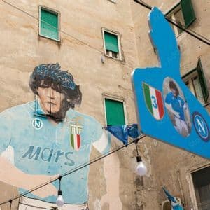 Un murale del calciatore Diego Maradona con una maglia azzurra su una facciata di un edificio, con luci pendenti e insegne davanti.
