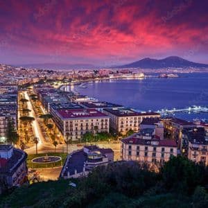 Veduta panoramica di una città costiera illuminata e della sua baia al tramonto, con una grande montagna sullo sfondo sotto un cielo drammatico e colorato.