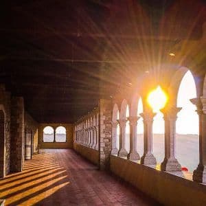 Il sole al tramonto splende attraverso gli archi di un colonnato in pietra, proiettando lunghe ombre dai pilastri su un pavimento piastrellato.