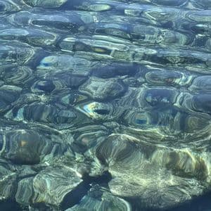 La luce del sole si riflette sulla superficie dell'acqua limpida e increspata, rivelando sul fondo pietre lisce e scure.