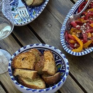 Una vista dall'alto di antipasti mediterranei, tra cui pane tostato, insalata di peperoni e melanzane grigliate, in ciotole di ceramica decorate su un tavolo di legno.