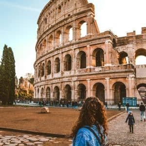 A woman with curly hair and a denim jacket stands on a cobblestone street, looking up at a large, ancient stone amphitheater under a clear sky.